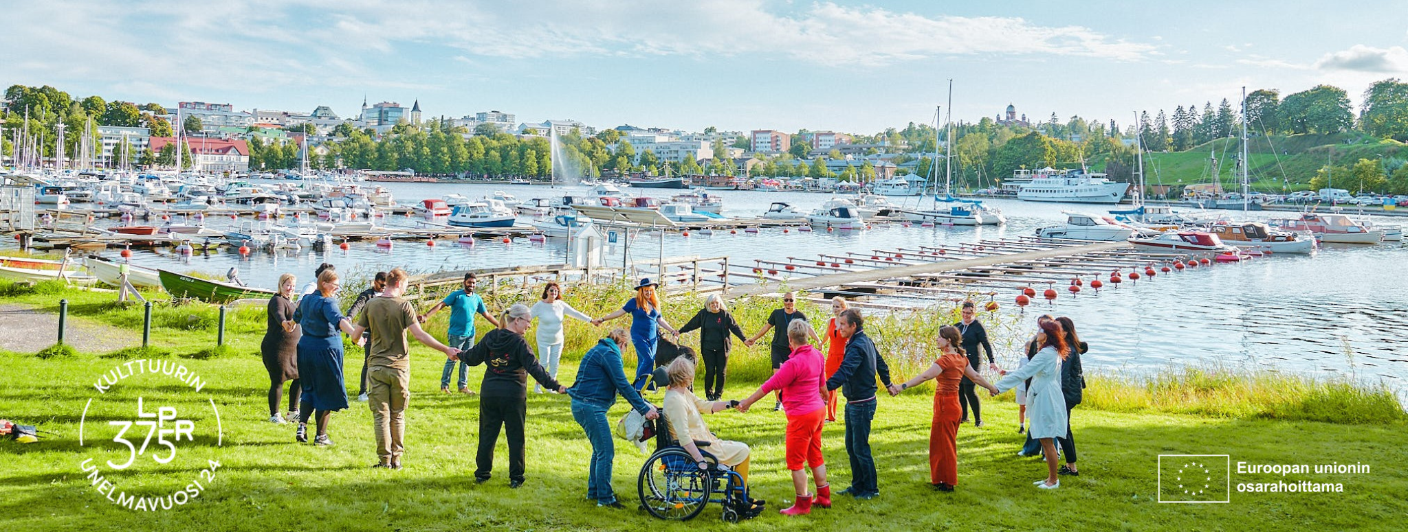 värikkäisiin vaatteisiin pukeutuneita ihmisiä piirissä Halkosaaressa. Taustalla Lappeenrannan satama.