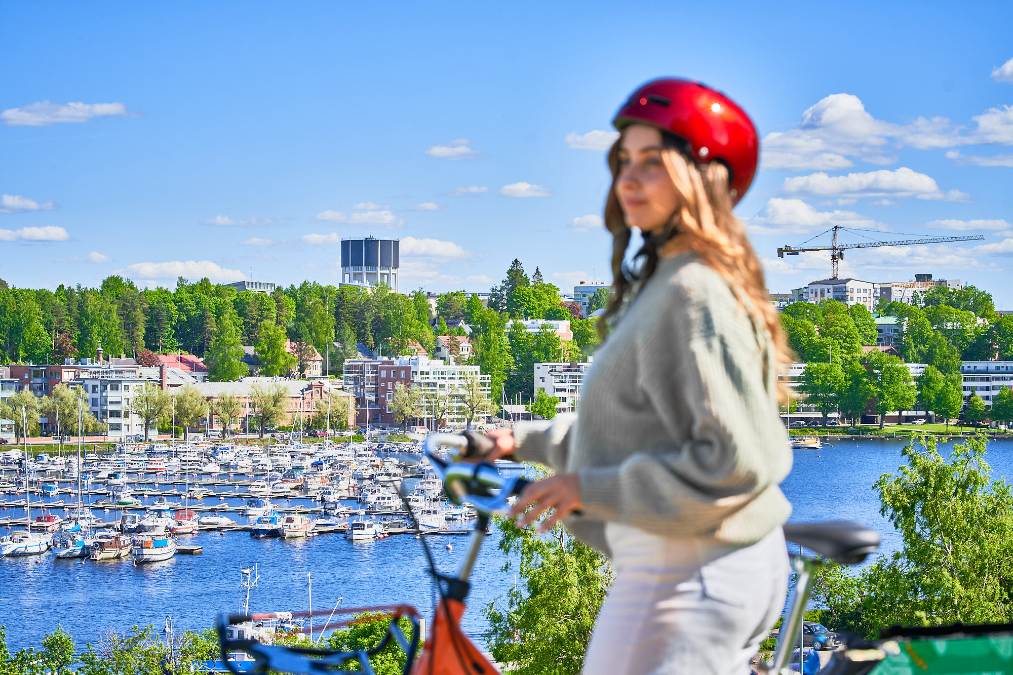 Kuvan etualalla on pyöräilijä aurinkoisena päivänä. Taustalla näkyy Lappeenrannan Kaupunginlahti, vihreitä puita ja taloja.