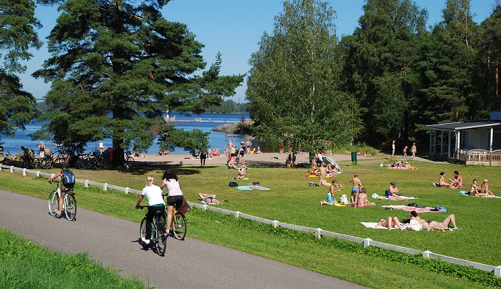 Myllysaaren pikniknurmella on paljon ihmisiä helteisenä päivänä, pyörätiellä kulkee pyöräilijöitä