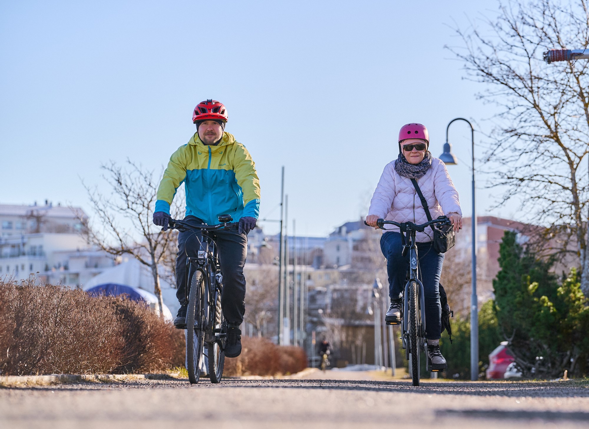 Pyörätien tasolta kuva miehestä ja naisesta pyöräilemässä. Taustalla valopylväitä, puita, pensaita, kaupungin rakennuksia.