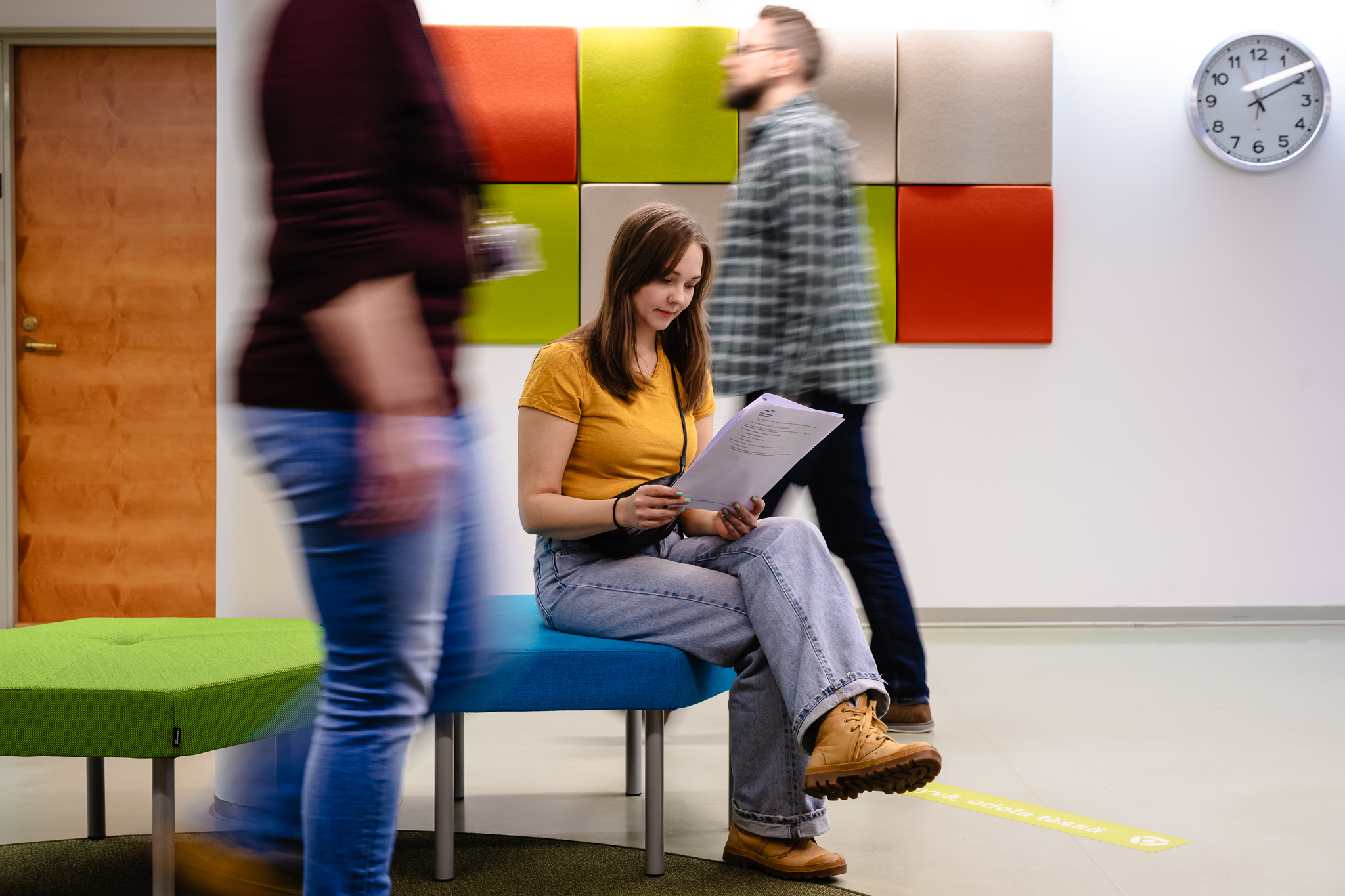 Naishenkilö lukee papereita istuen sisätilassa. Hänen ohitseen kulkee ihmisiä.