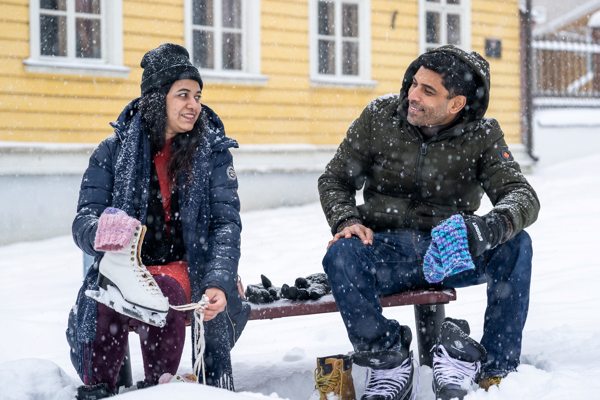 Kaksi henkilöä pukee luistimia lumisateessa penkillä istuen.