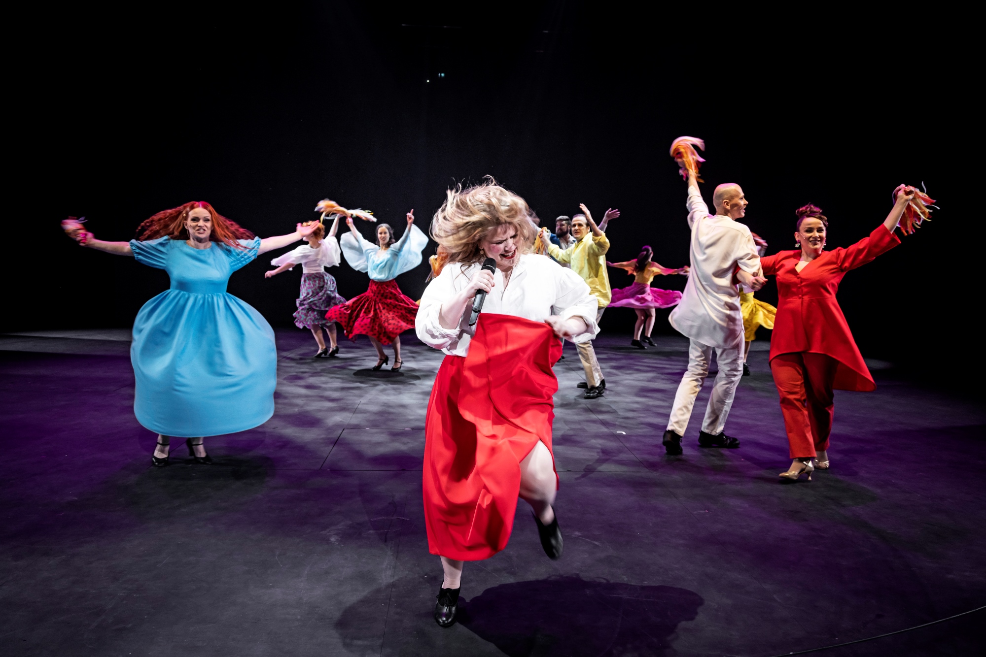 Ihmisiä tanssimassa värikkäissä vaatteissa teatteri lavalla.