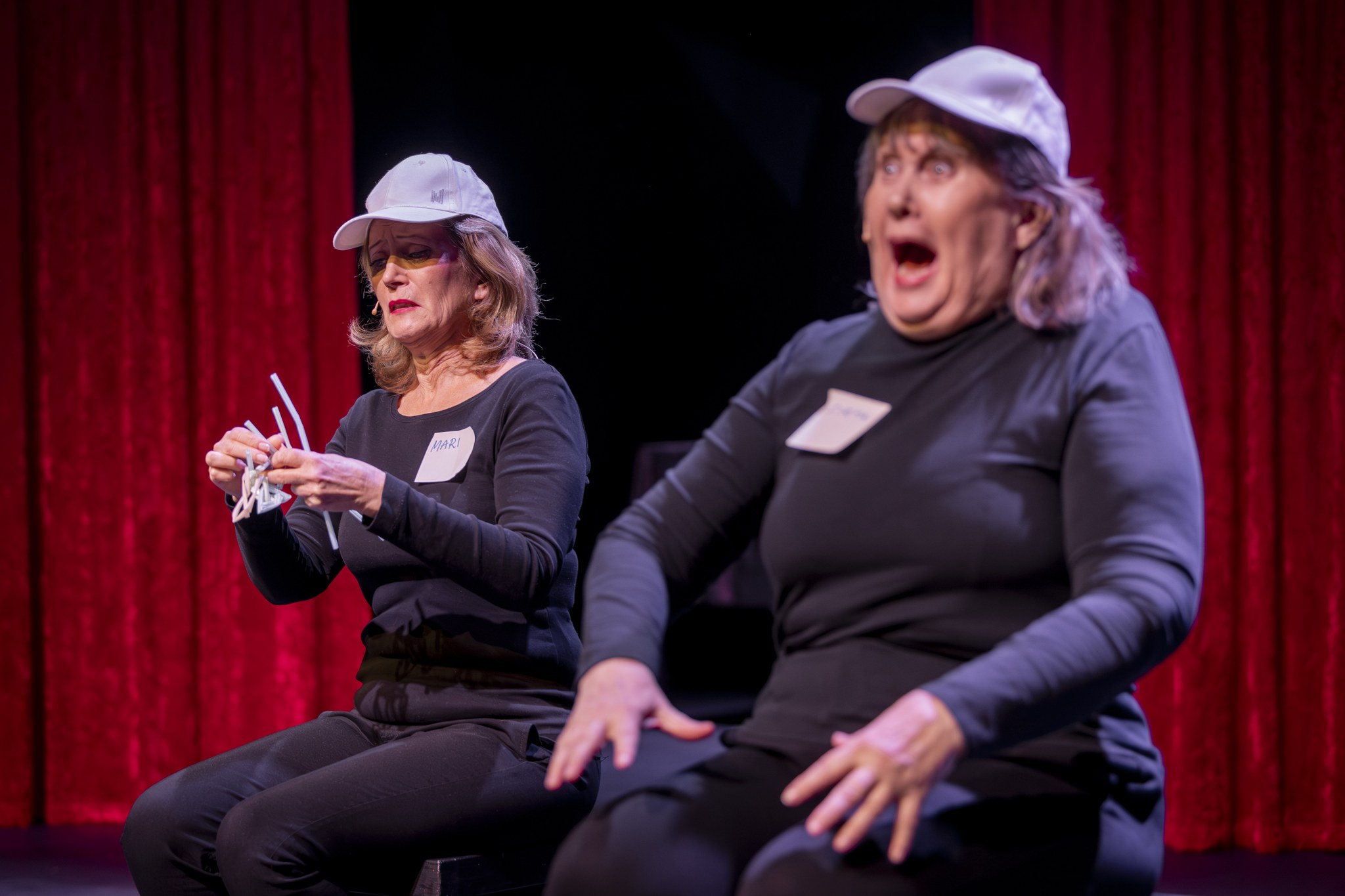 Kaksi vanhempaa naista istuu tuoleilla vierekkäin. Toisella on kasvoillaan ahdistunut ja epätoivoinen ilme, toinen huutaa yllättyneenä. 