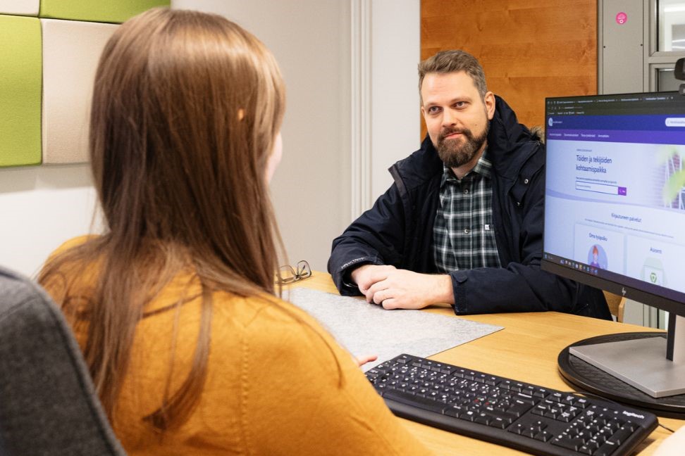 Henkilöt istuvat toimistopöydän ääressä toisiaan vastapäätä ja tietokoneen näytöllä näkyy Työmarkkinatorin etusivu.