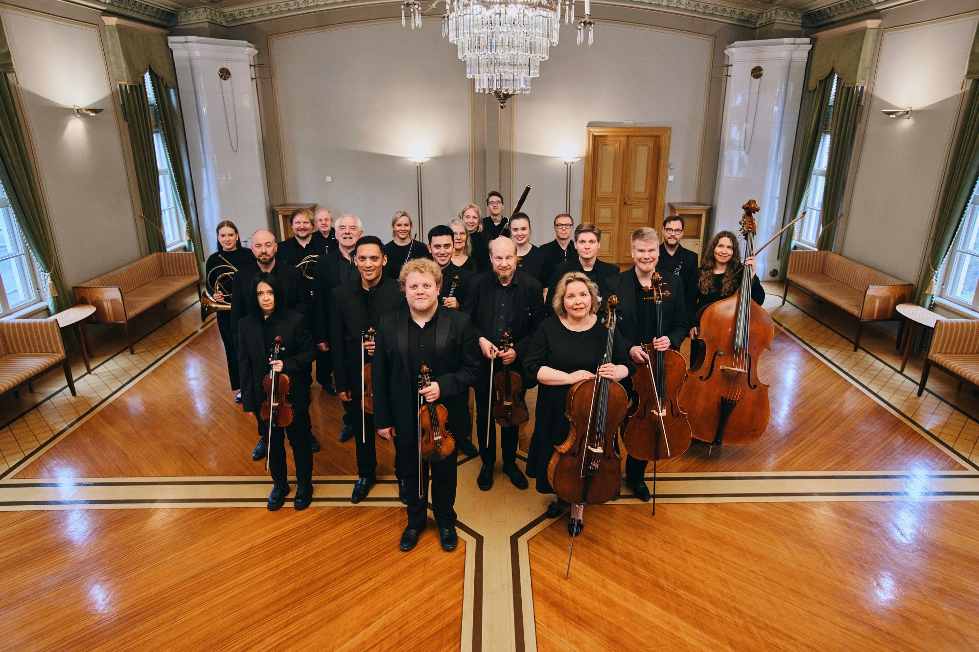 Lappeenrannan kaupunginorkesterin muusikoiden ryhmäkuva. Muusikot seisovat juhlavassa salissa mustiin pukeutuneina ja soittimet kädessä.