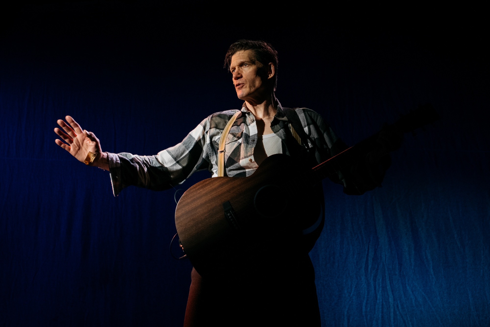 Ruutupaitaan ja henkseleihin pukeutunut mies seisoo kitara kädessä dramaattisessa valossa. 