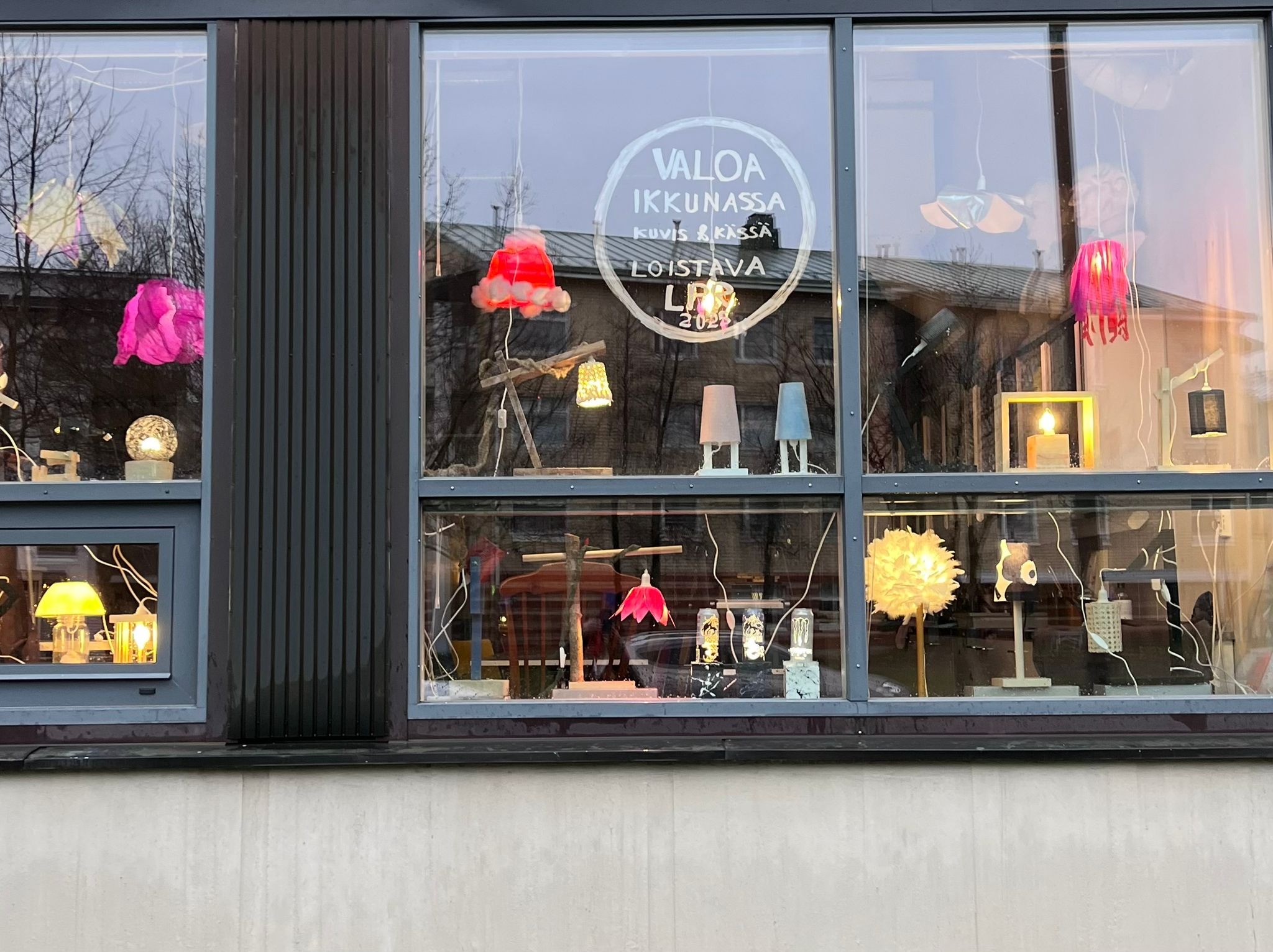 Erilaisia omatekoisia valaisimia asetettu ikkunaan näytille, ikkunassa ympyrän sisällä teksti Valoa ikkunassa, kuvis & kässä, Loistava LPR 2025.