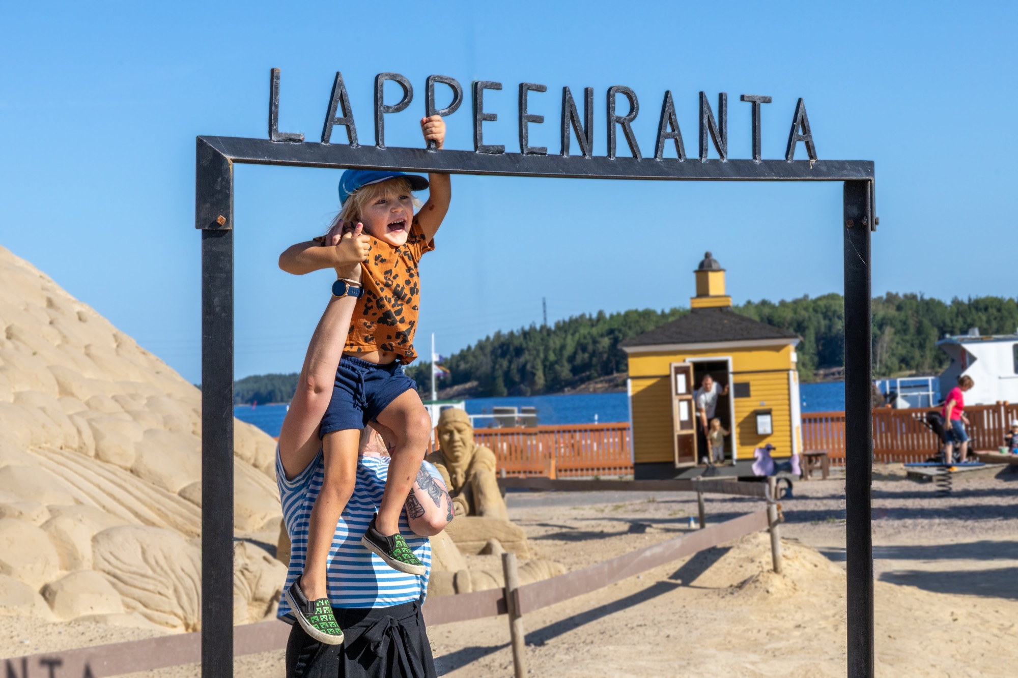 Nainen nostaa pientä poikaa roikkumaan porttiin jonka päällä on Lappeenranta-kirjaimet. Taustalla näkyy hiekkalinnaa ja Saimaa.