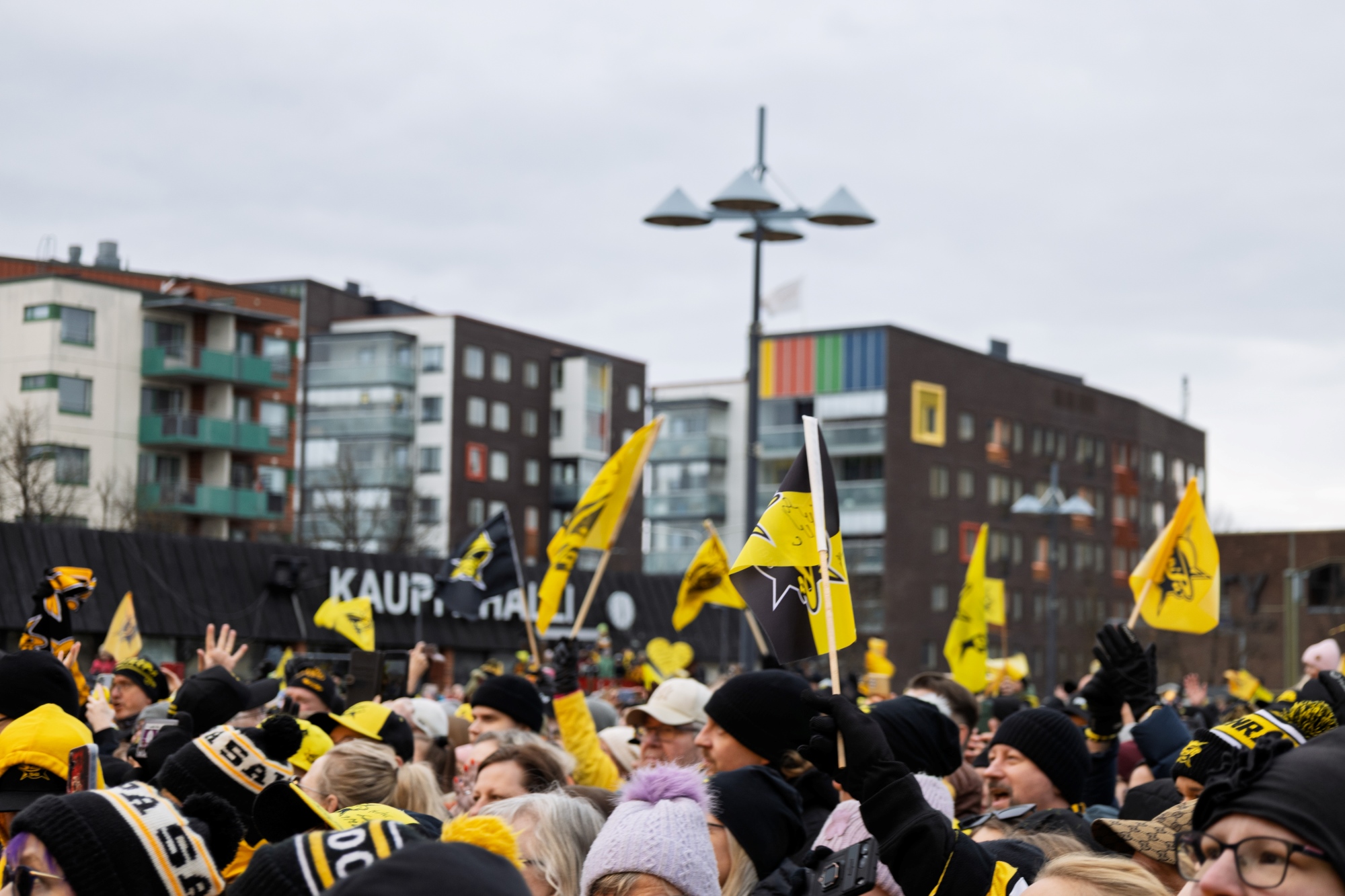 SaiPa-jääkiekkojoukkueen kannatusasuihin pukeutuneita ihmisiä kokoontuneena torille heiluttamaan lippuja. Taustalla kerrostaloja.