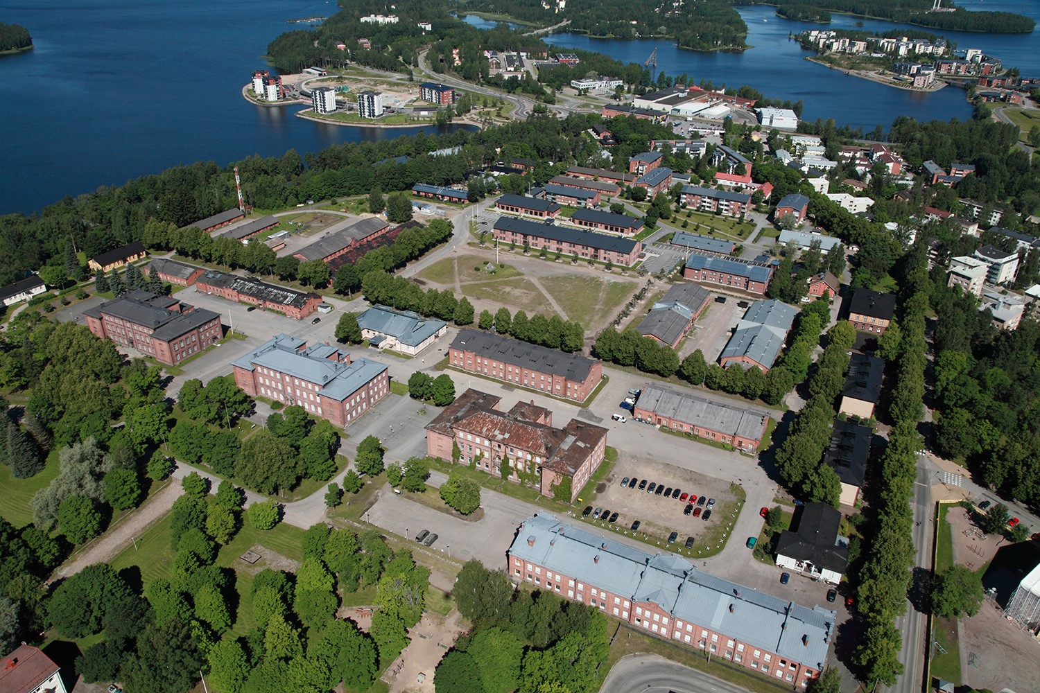 Ilmakuva, jossa näkyy punatiilisiä rakennuksia, puita ja aukio. 