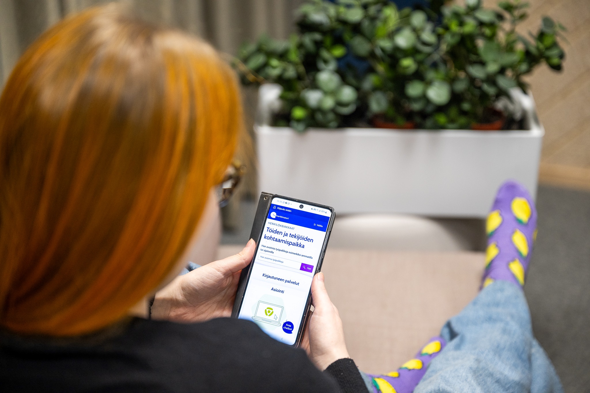 Henkilö katsoo kännykästä tietoja Työmarkkinatorilta. Kuva on otettu henkilön olkapään takaa ja hän istuu sohvalla sisätilassa.