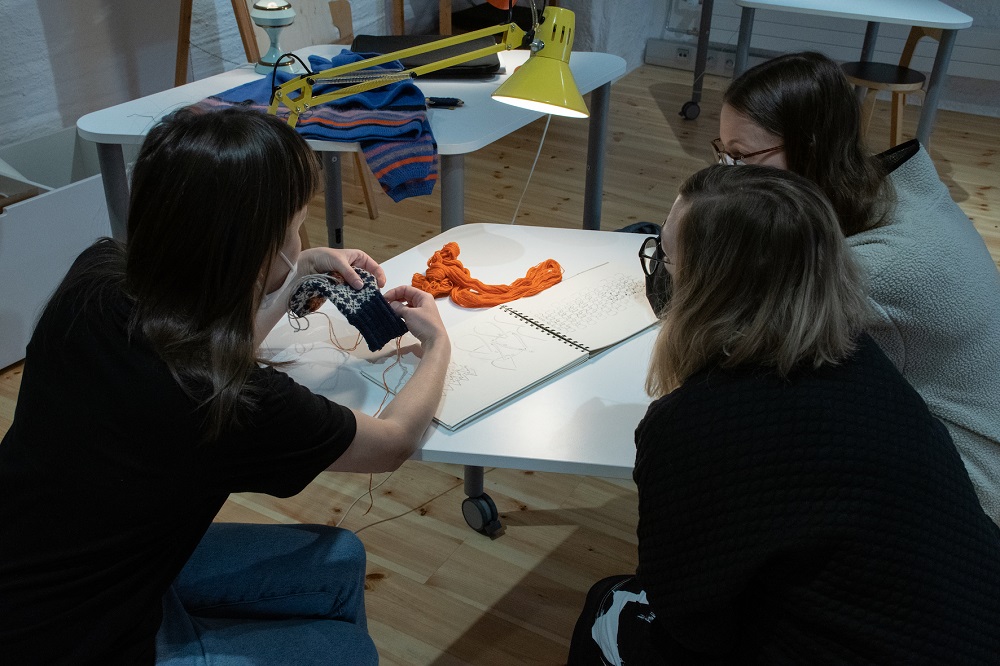 Kolme henkilöä työskentelee neulomuksen parissa kumartuneina salmiakinmuotoisen pöydän ylle. Työskentelyvalo on kohdistettu käsityöhön.