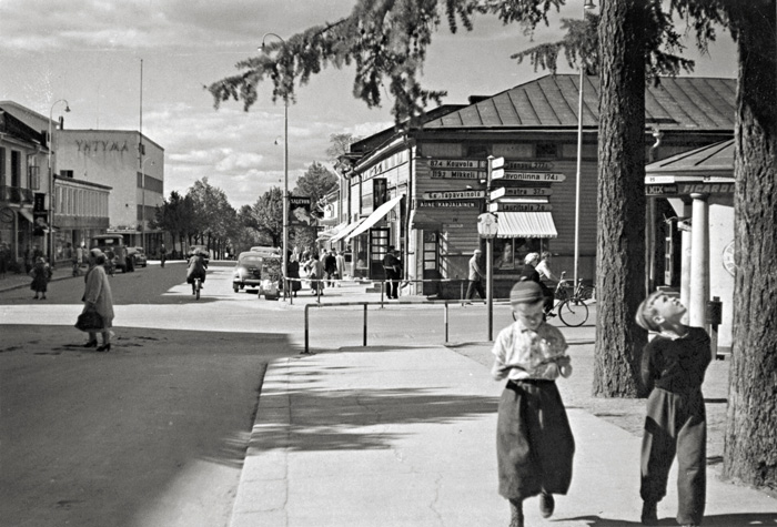 Lappeenrannan Kauppakadun ja Valtakadun risteys 1950-luvulla, etualalla oikealla kaksi poikaa kävelee jalkakäytävällä. Taustalla näkyy vanhoja liikerakennuksia.