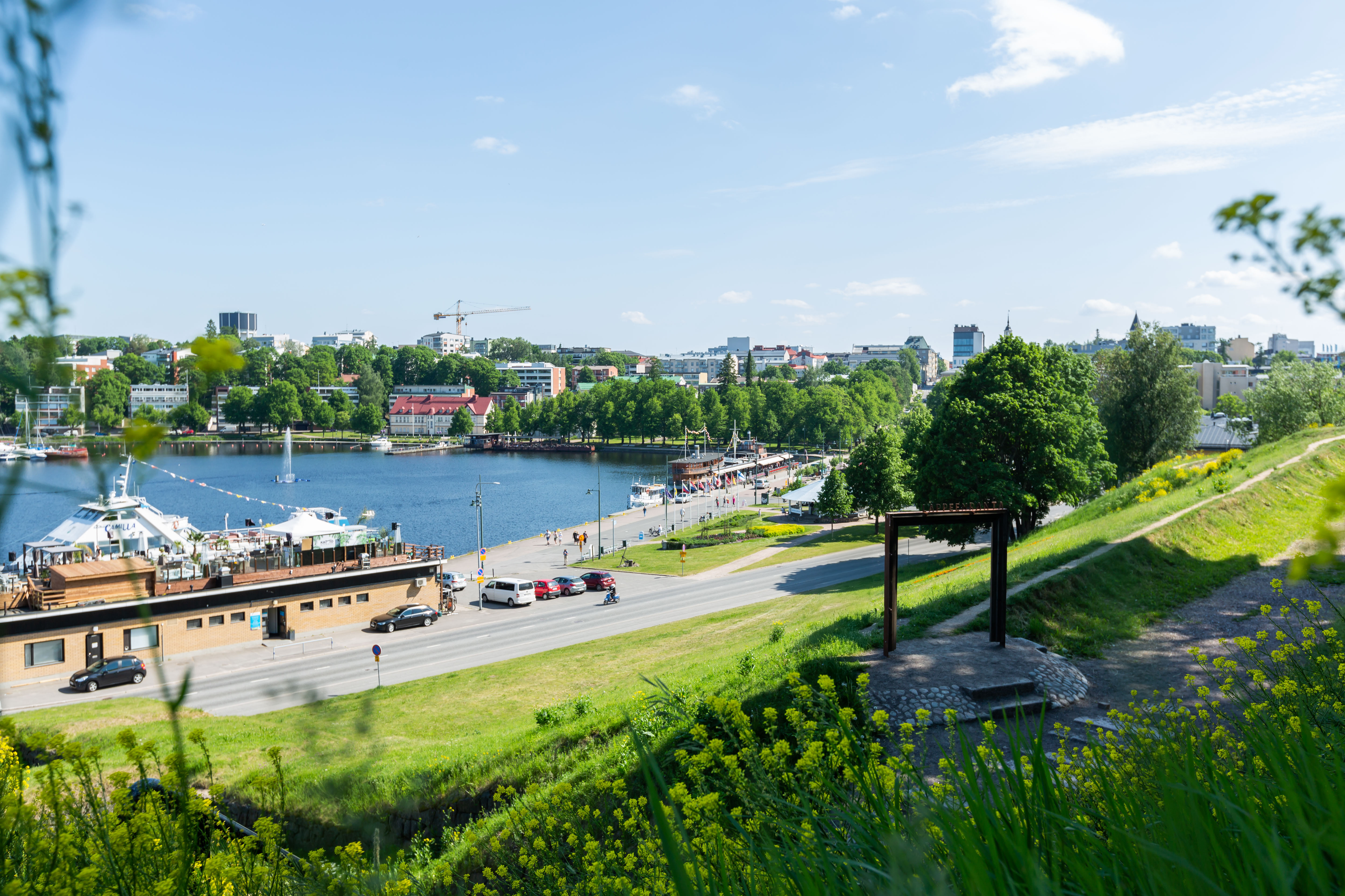 Vehreä kuva Lappeenrannan Satamasta kuvattuna linnoituksen valleilta päin. Taustalla näkyy sininen Saimaa ja laivaravintoloita. 